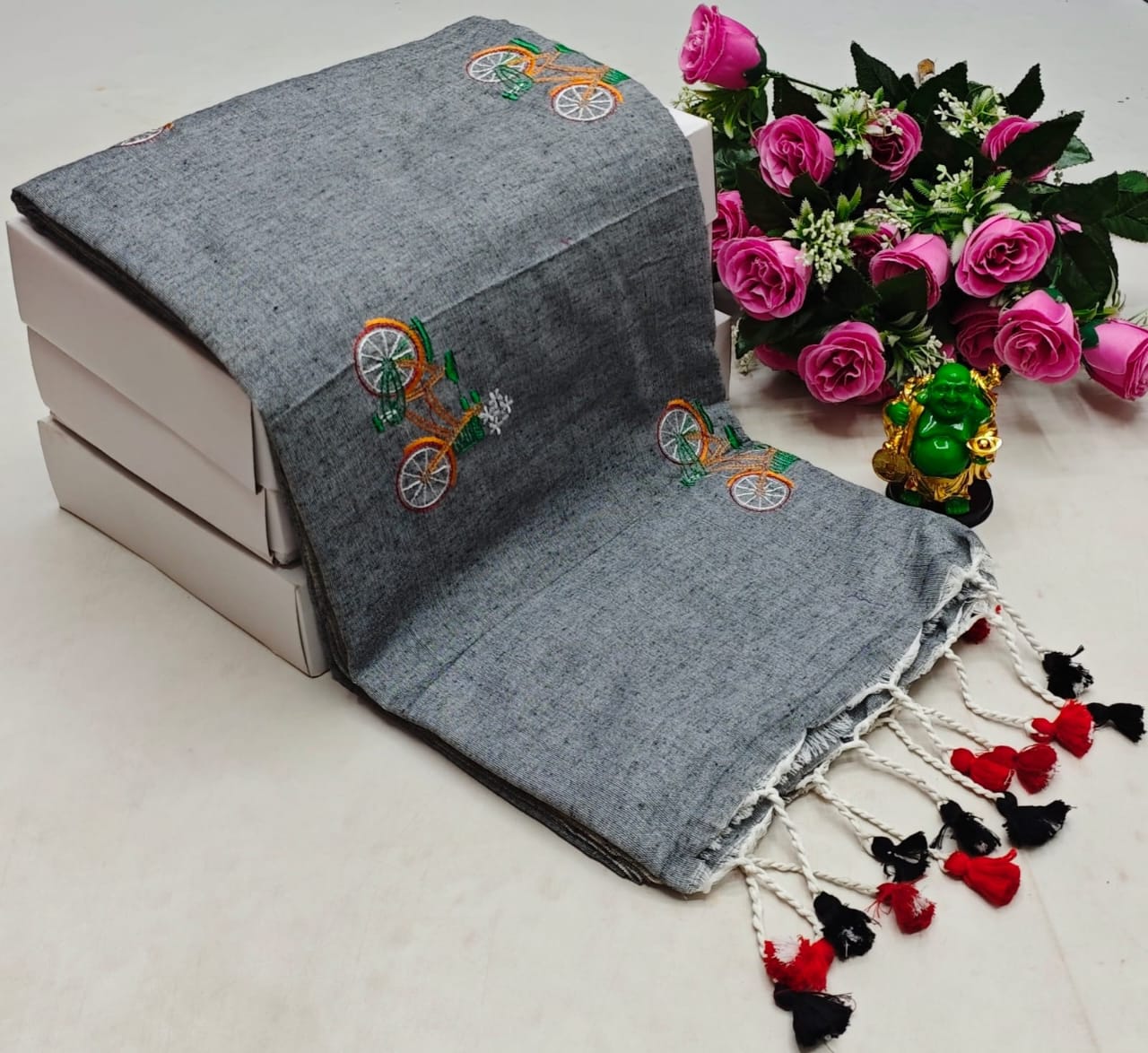 Gray scarf with embroidered designs and tassels on a white surface with flowers in the background